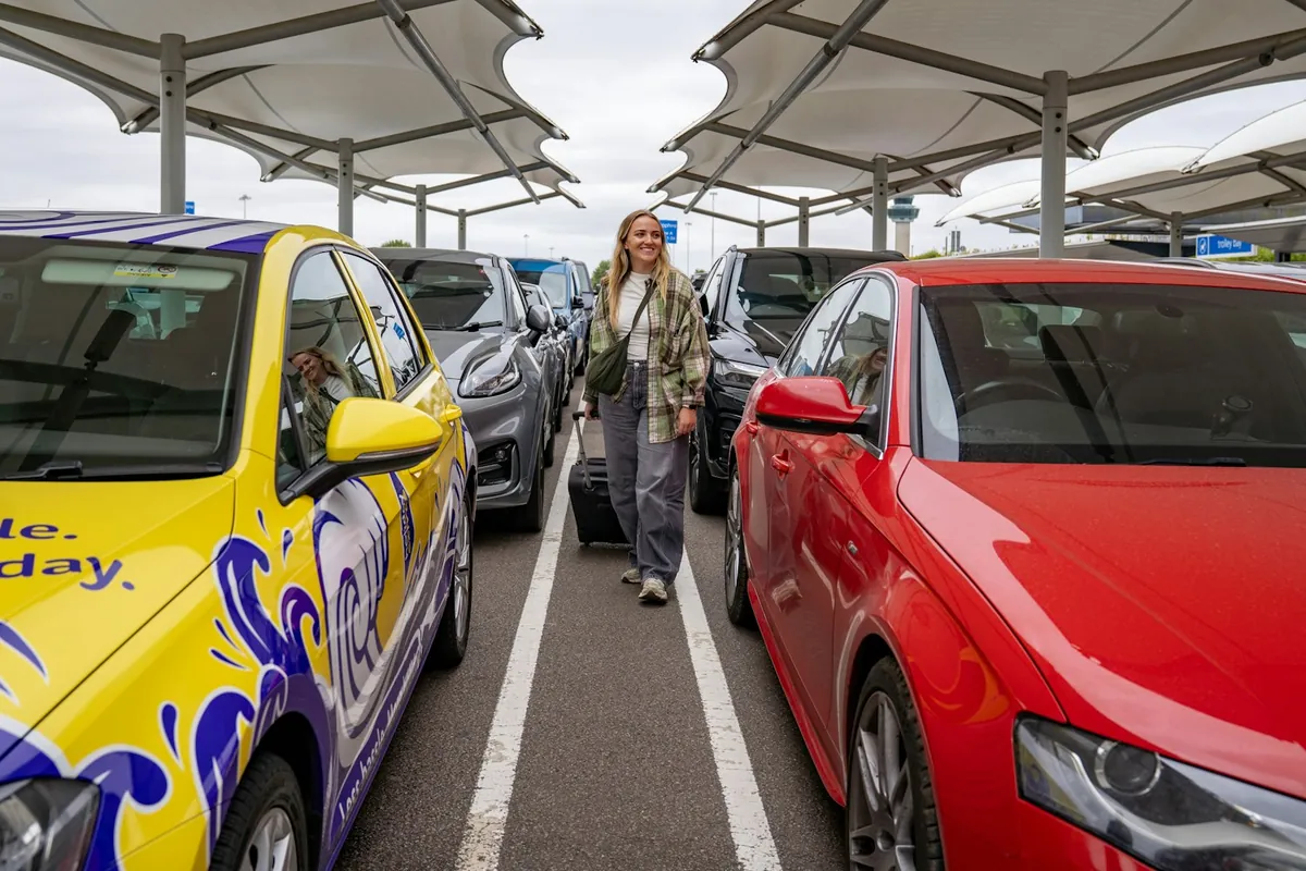 Reisender mit Gepäck geht zwischen Mietwagen auf einem Flughafenparkplatz