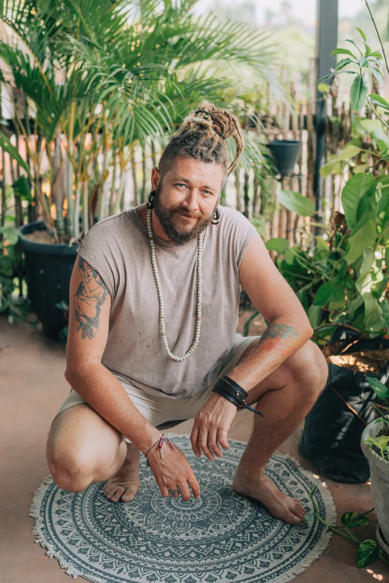 Chenzo, founder of NomadsToolsLab, crouching on a round rug in a garden full of plants