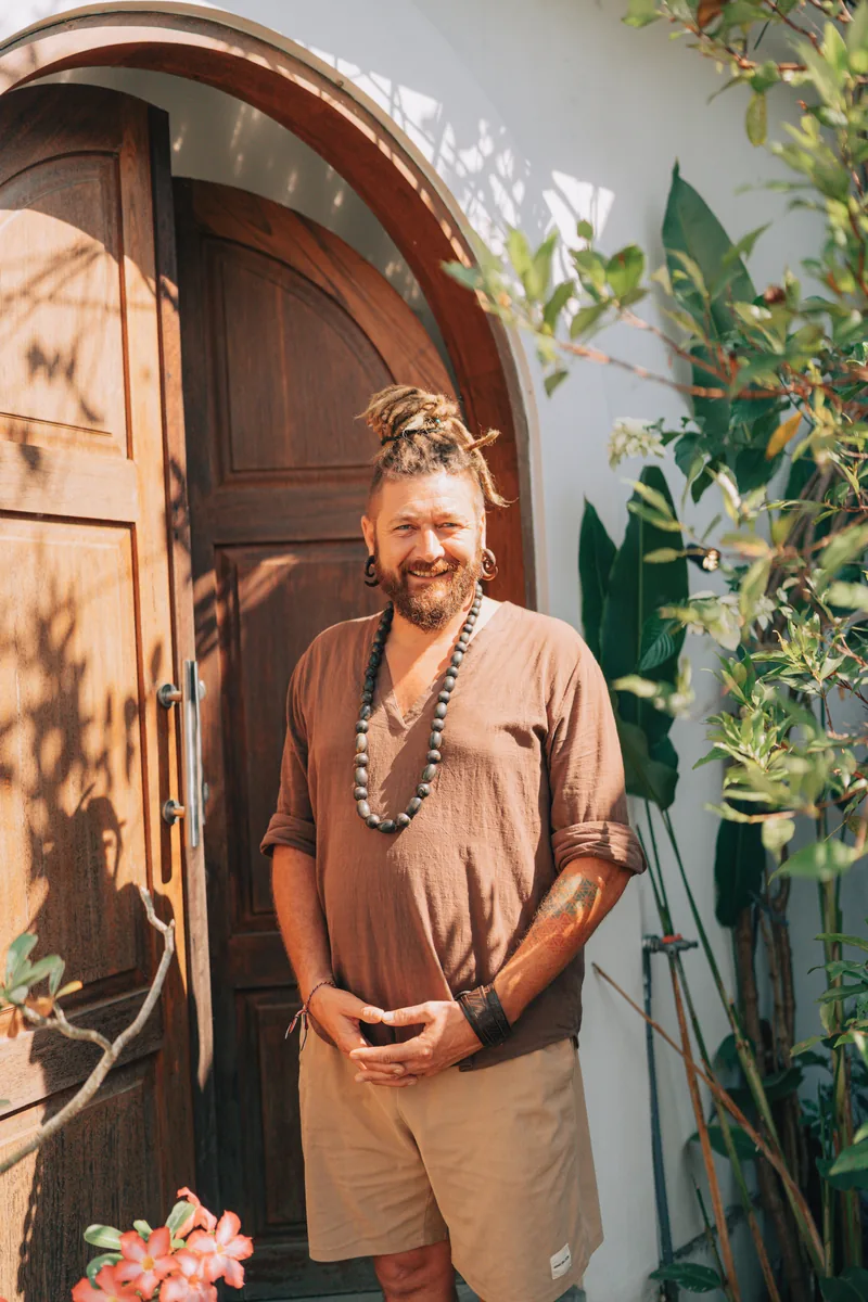 Chenzo, fundador de NomadsToolsLab, de pie junto a una puerta de madera con plantas y flores rosas