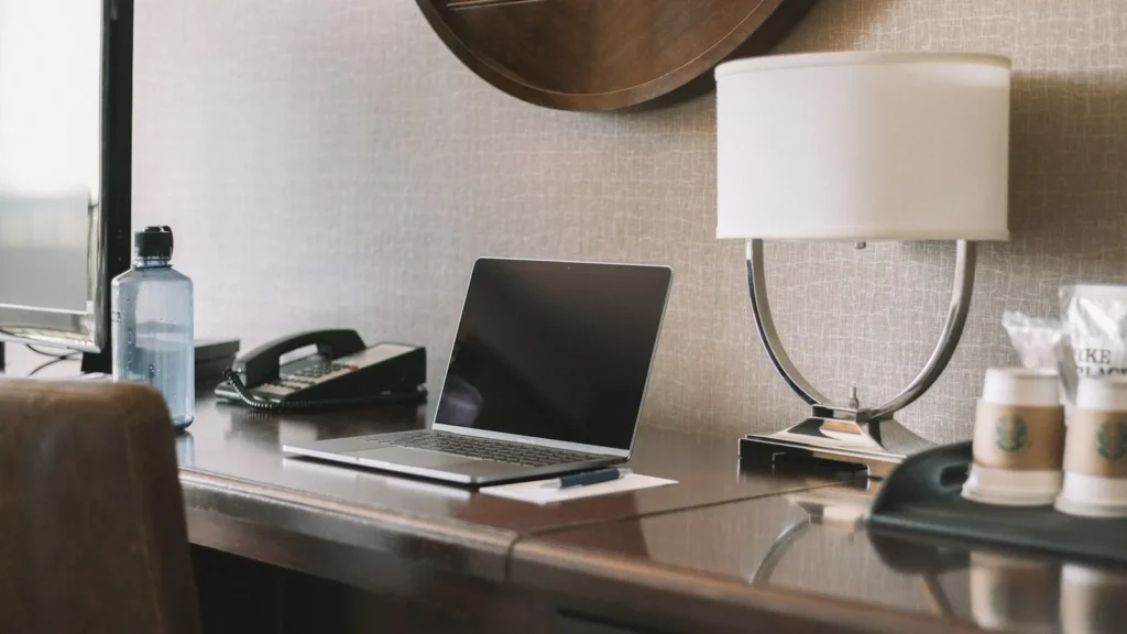 Laptop on a hotel room desk next to a lamp and coffee
