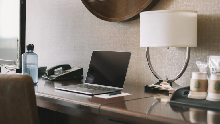Laptop on a hotel room desk next to a lamp and coffee