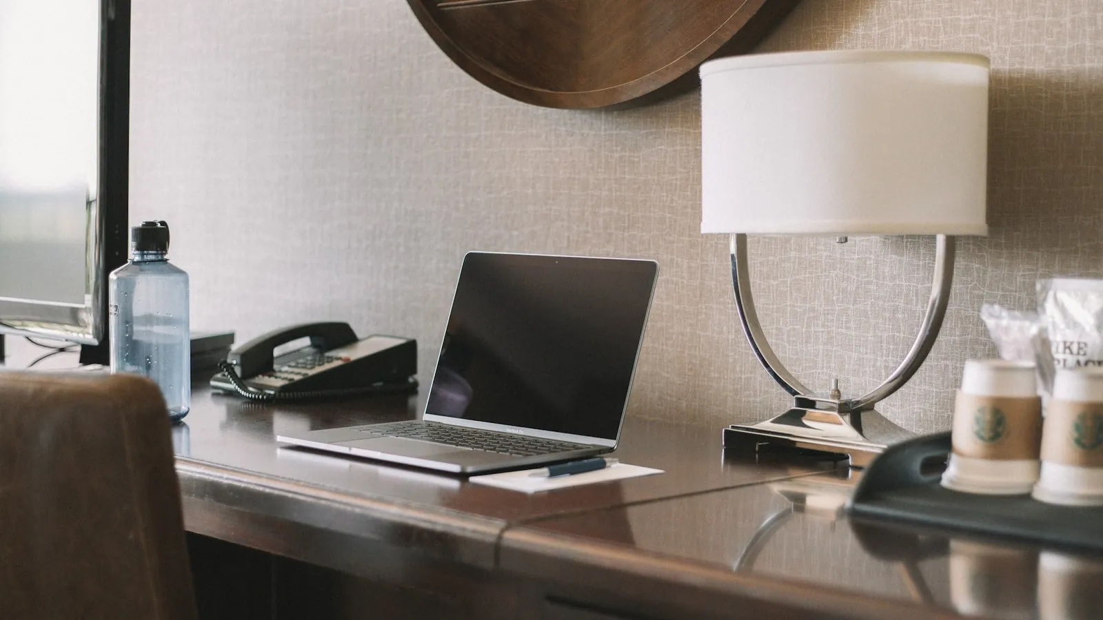 Laptop on a hotel room desk next to a lamp and coffee