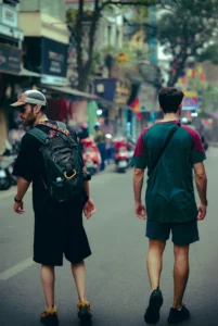 Two backpackers with daypacks walking through a narrow Hanoi Old Quarter street lined with shophouses