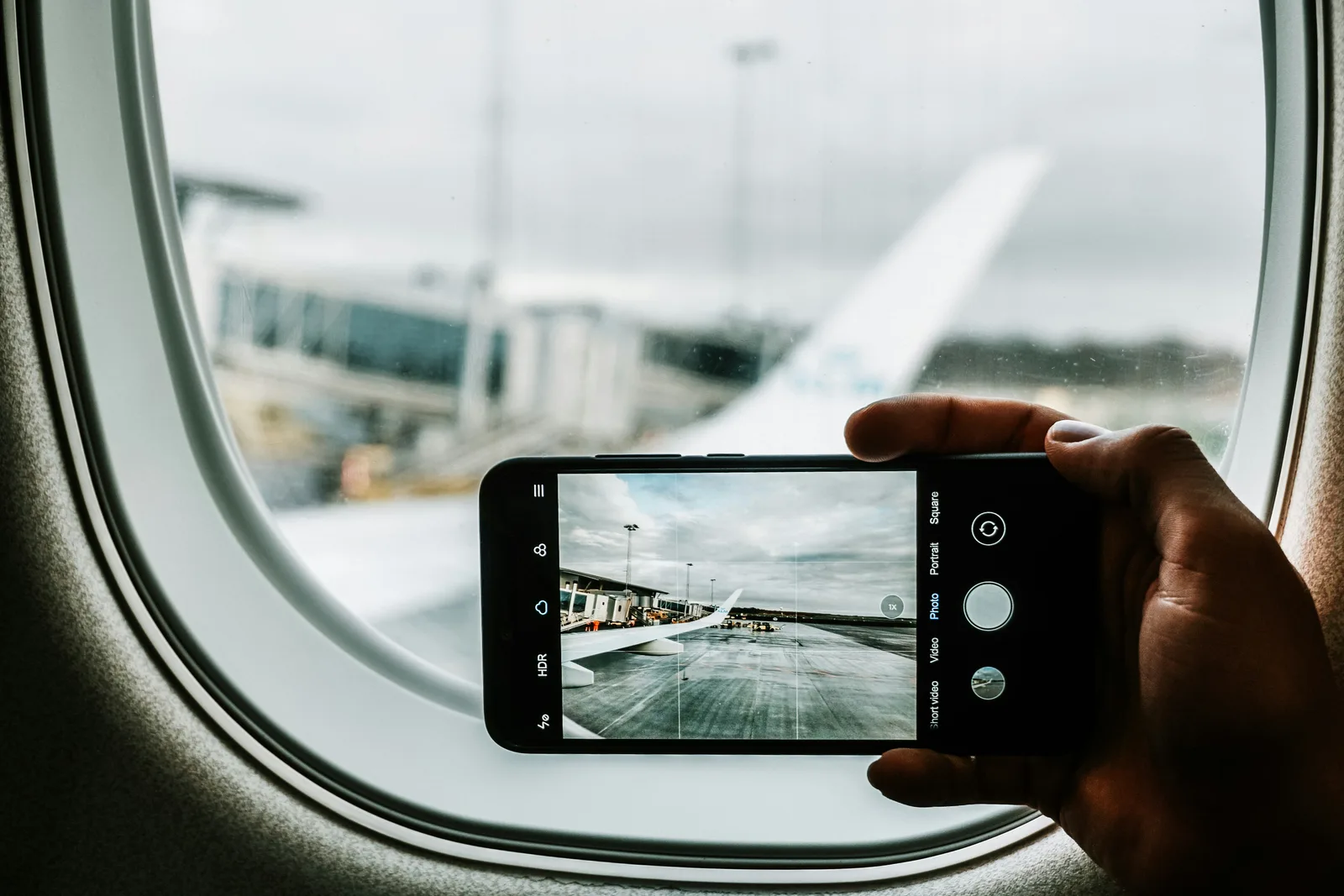 Hand hält ein Handy und fotografiert eine Flugzeugtragfläche durch das Fenster