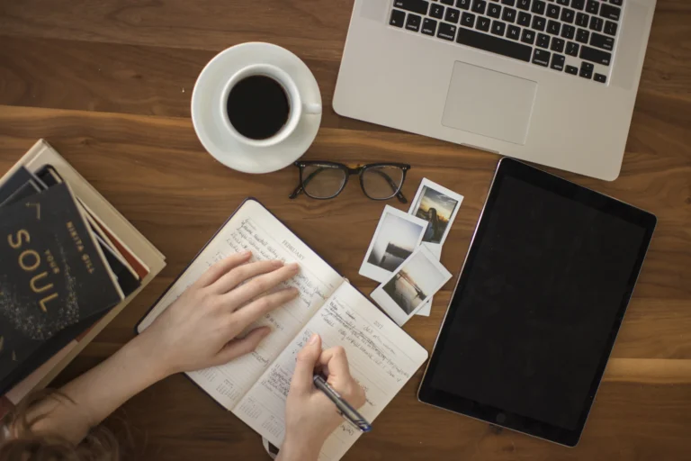 Person writing in a notebook next to a laptop and a cup of coffee