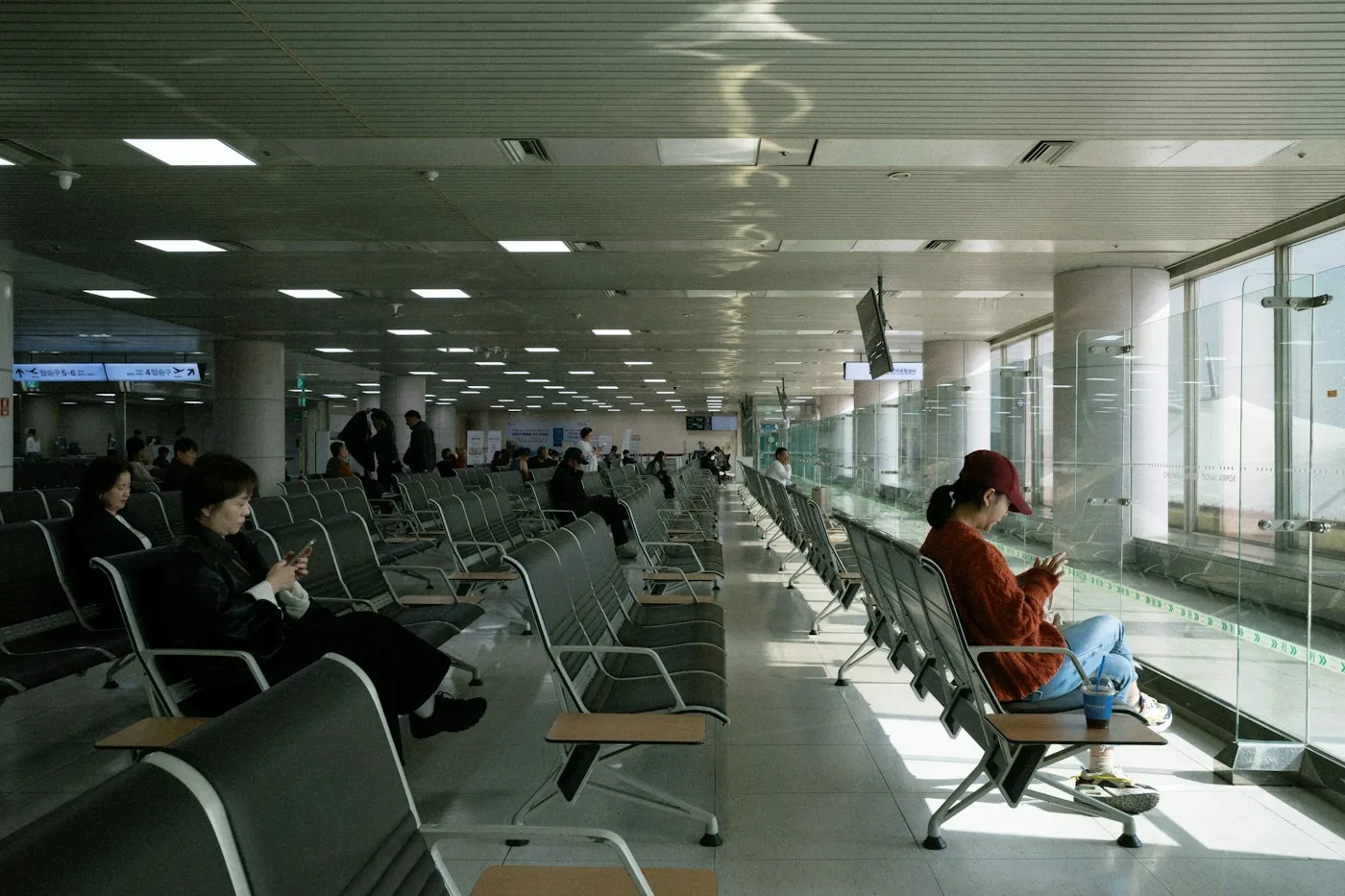 Travelers sitting on rows of seats in a long airport lounge with large windows, most of them on their phones