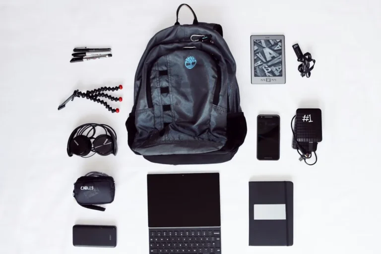 A flat lay of a black backpack, laptop, headphones, phone and other travel gear on white