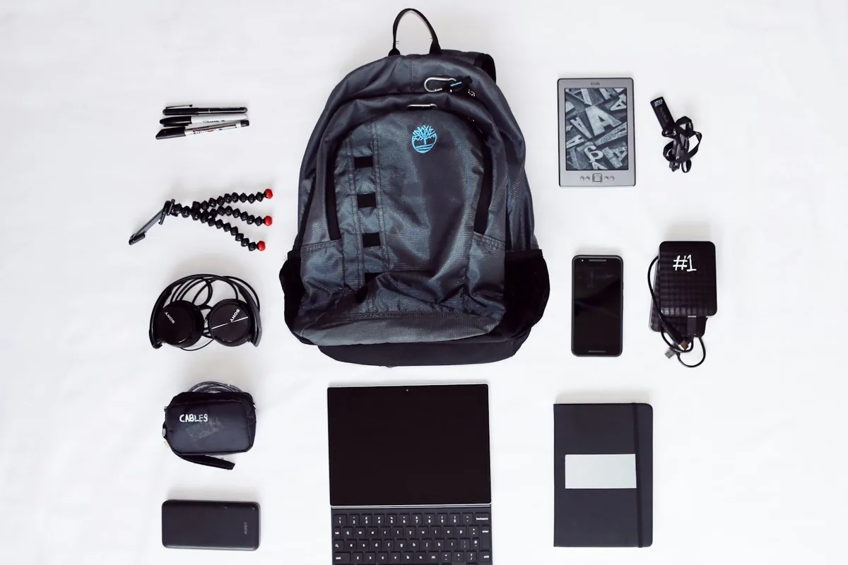 A flat lay of a black backpack, laptop, headphones, phone and other travel gear on white