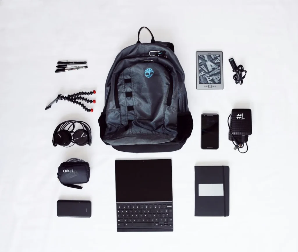 Flat lay of a backpack, headphones, smartphone, tablet with keyboard and other travel gear on a white background