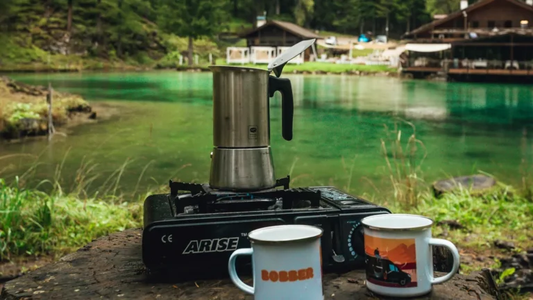 Moka pot on a camping stove next to two mugs by a lake, best coffee maker for remote workers