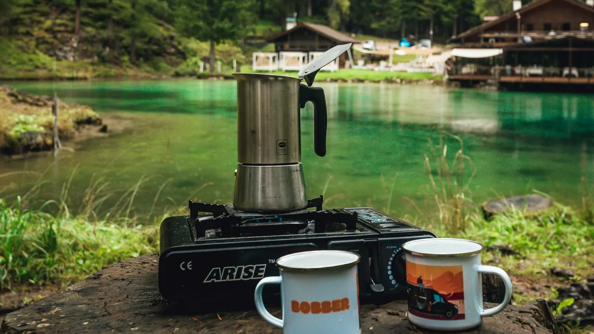 Moka pot on a camping stove next to two mugs by a lake, best coffee maker for remote workers