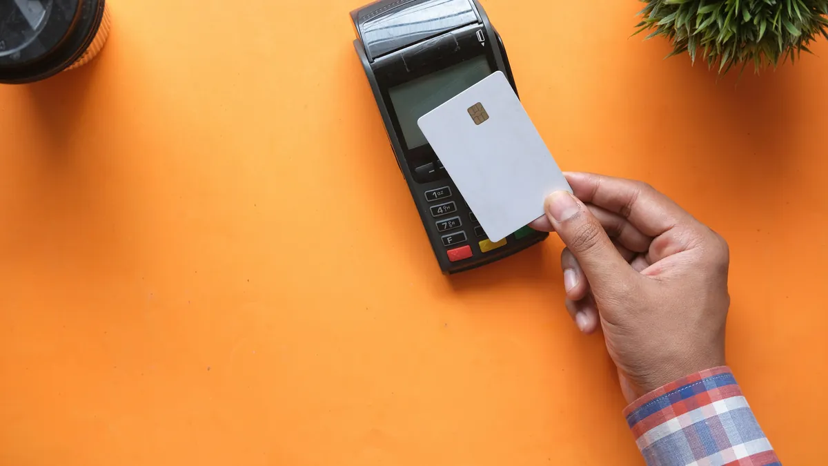 Hand holding a blank bank card to a payment terminal on an orange table, best travel credit cards