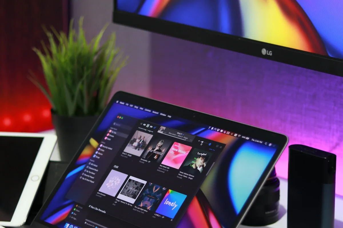 Black laptop on a desk under a larger monitor with a tablet and a black power bank beside it