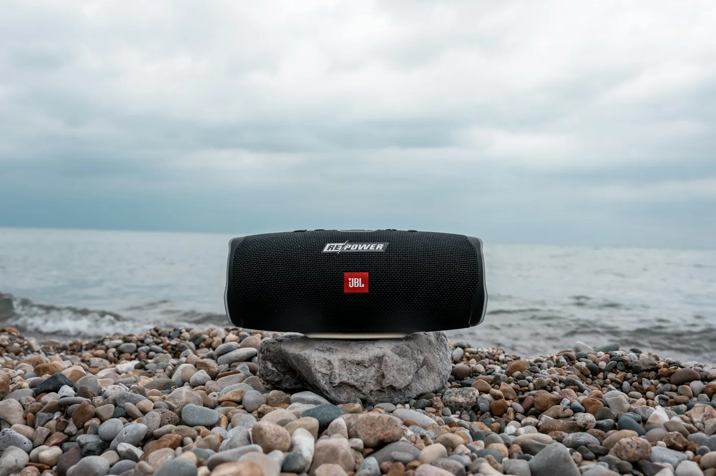 A black portable Bluetooth speaker on a flat rock on a pebble beach by the sea