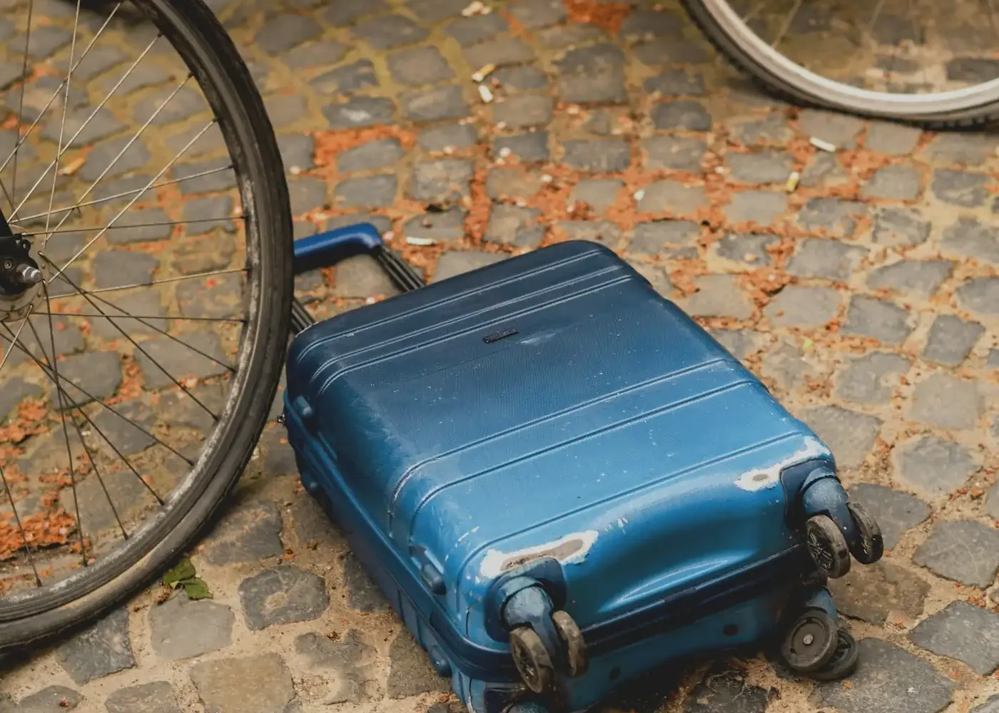 Blue hardside suitcase lying on a cobblestone street next to bicycle wheels, with visible scuffs