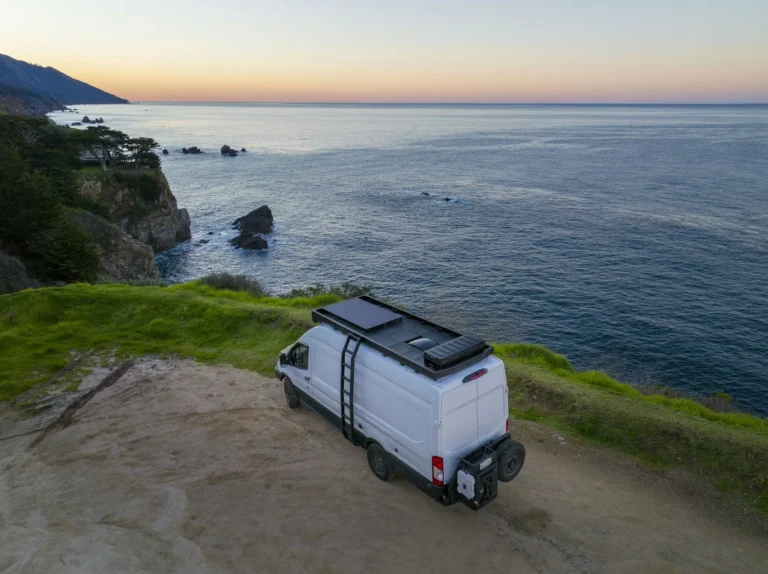 White van with solar panels on its roof parked on a grassy cliff above the ocean at sunset