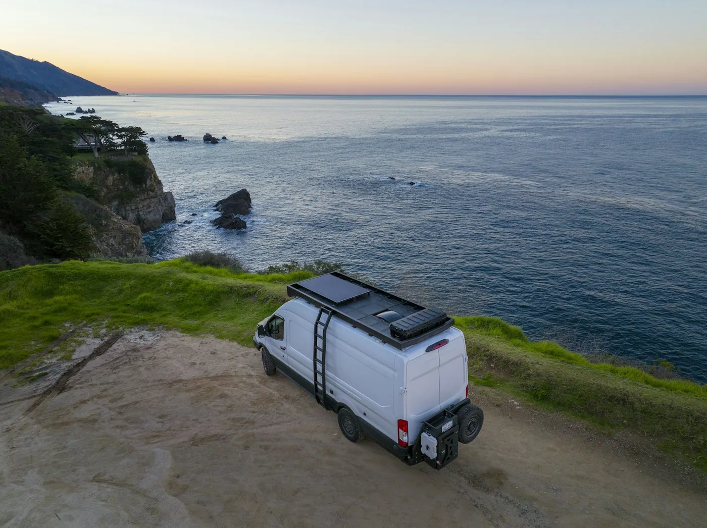 White van with solar panels on its roof parked on a grassy cliff above the ocean at sunset