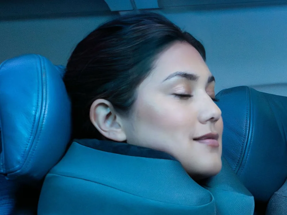 Woman with her eyes closed resting on a teal Cabeau Evolution S3 neck pillow in an airplane seat