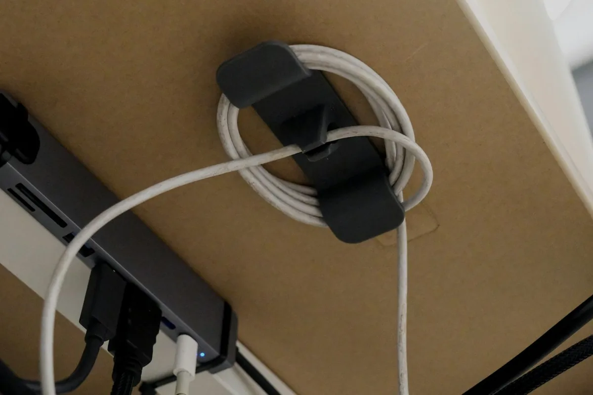 Een witte oplaadkabel, opgerold en vastgezet met een zwarte clip onder een bureau