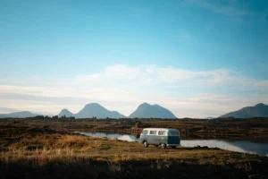 A vintage camper van parked by a calm lake with mountains behind
