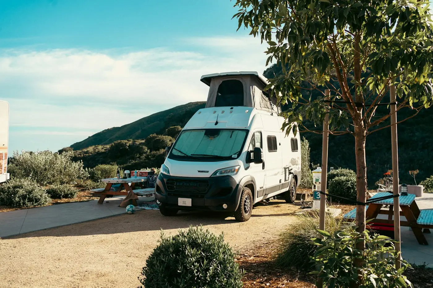 White camper van with a pop-up roof parked at a campsite with picnic tables, mountains behind it and a clear sky