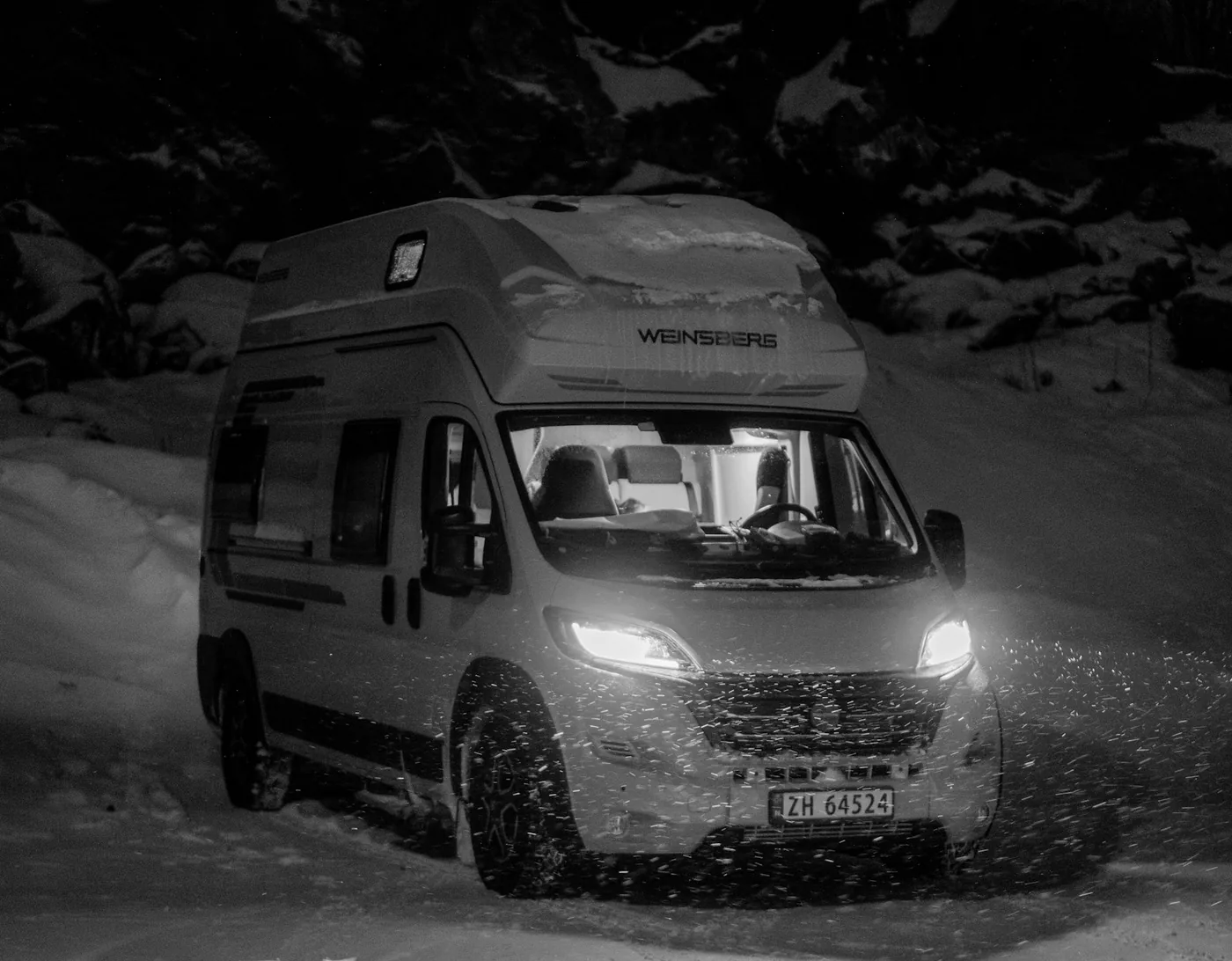 Camper van with its headlights on parked on a snowy slope at night in falling snow