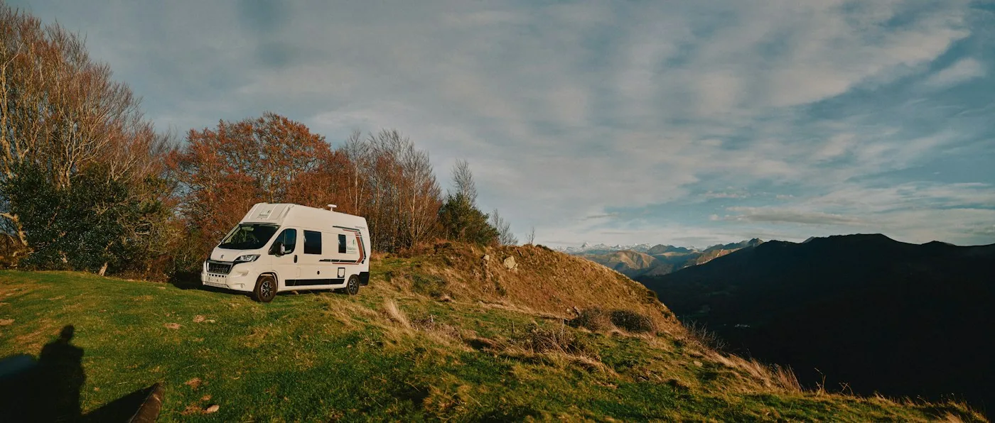 White camper van parked on a grassy hillside with autumn trees, mountains and a cloudy sky behind it