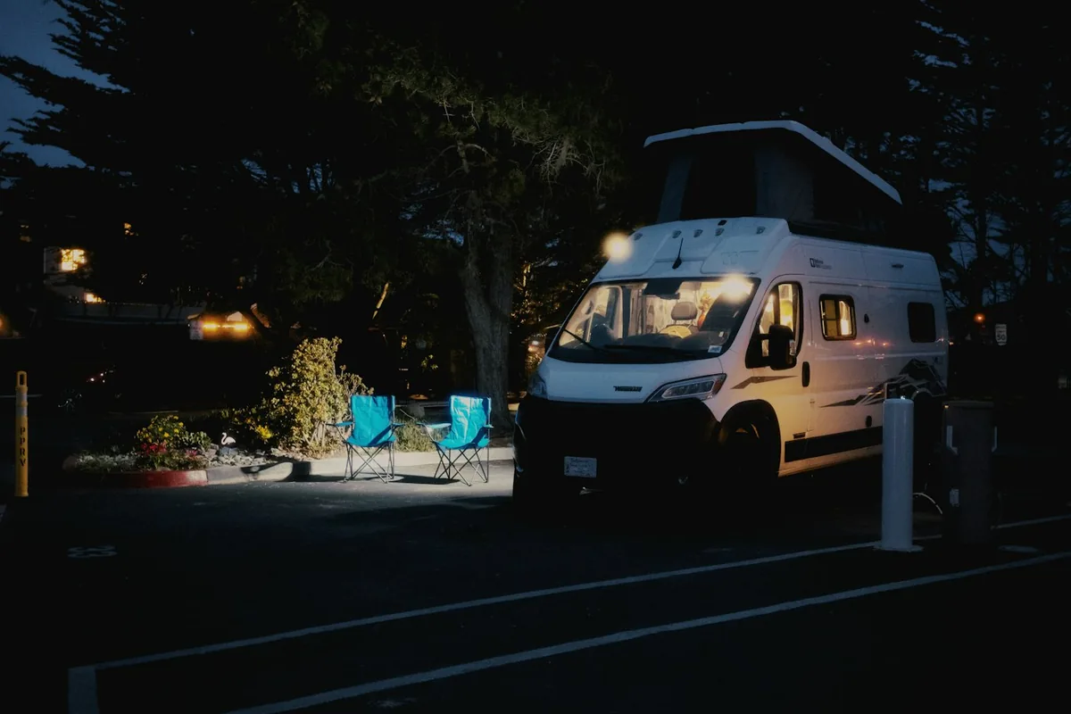 Campervan with its interior light on at a campsite at night, two camping chairs beside it