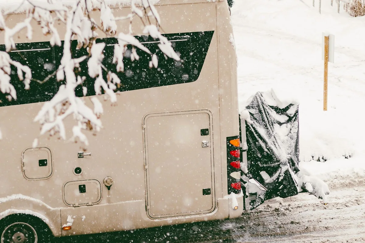 Beiger Camper auf einer verschneiten Straße, Schnee fällt, mit einer schwarzen Abdeckung über dem Fahrradträger am Heck