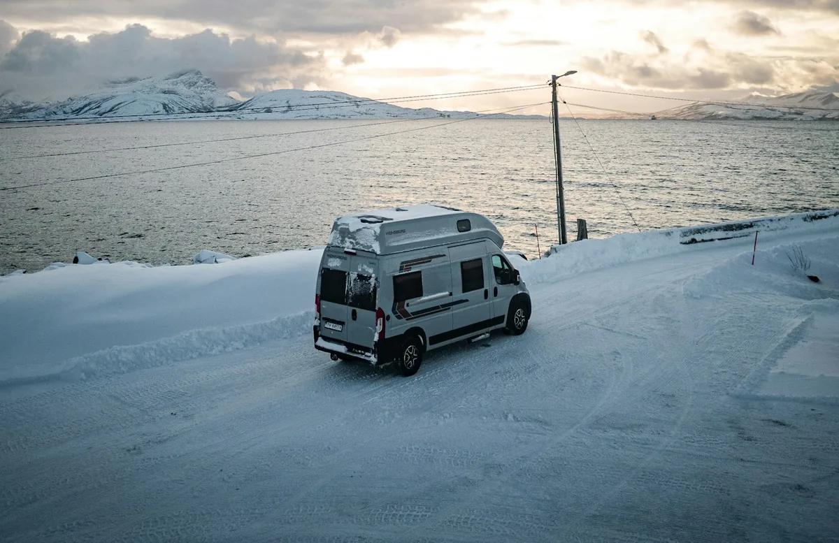 Camper parkt bei Sonnenuntergang auf einer verschneiten Straße neben einem Fjord, ein kalter Ort zum Laden einer Lithiumbatterie