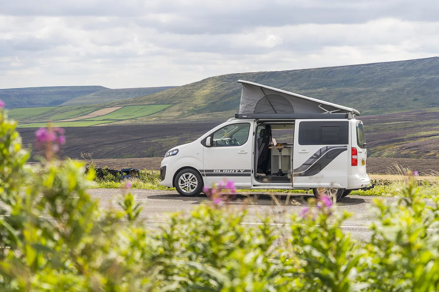 Camper van blanca con techo elevable y puerta lateral abierta aparcada al borde de una carretera en una zona de colinas