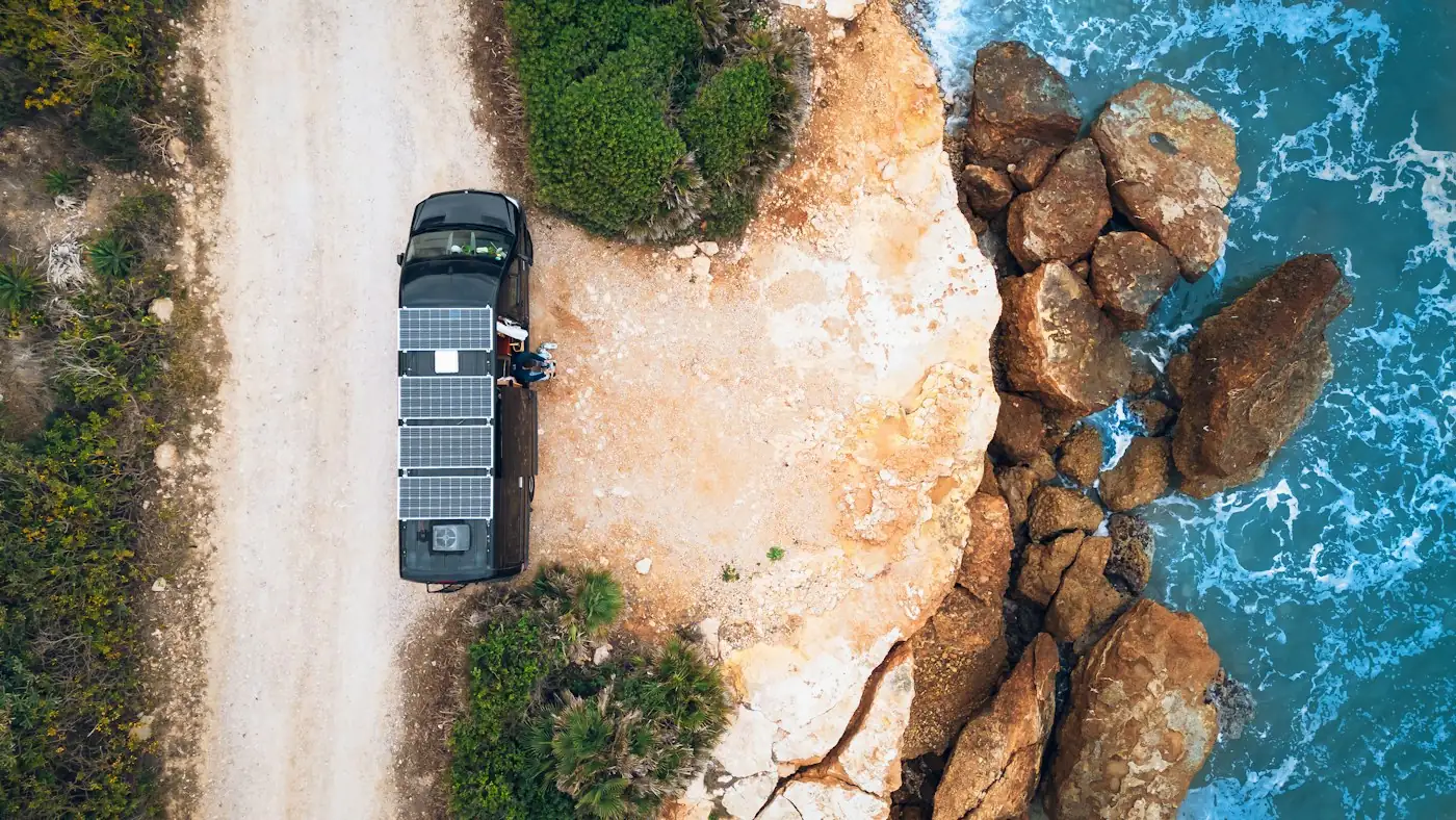 Vista aérea de una furgoneta camper con paneles solares en el techo, aparcada en un camino de tierra junto a una costa rocosa
