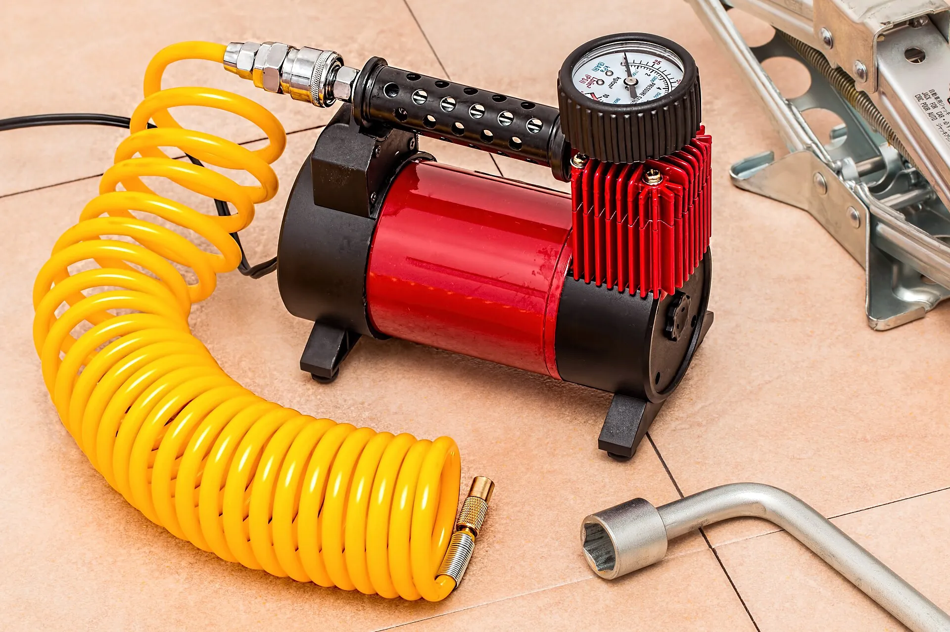 Red 12 V tire compressor with a pressure gauge and yellow coiled hose, next to a wheel brace and a jack