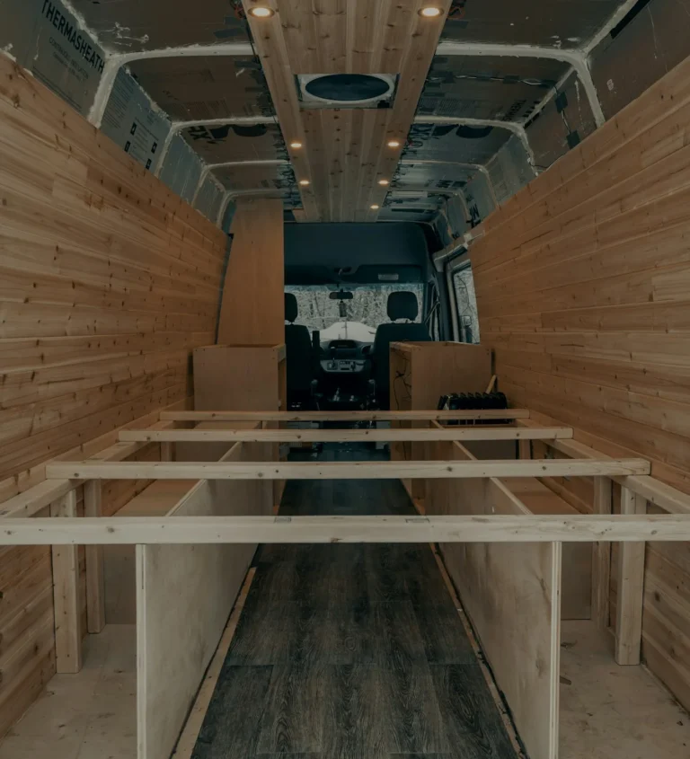 Inside a cargo van with wood paneling on the walls and ceiling and wooden floor framing in place