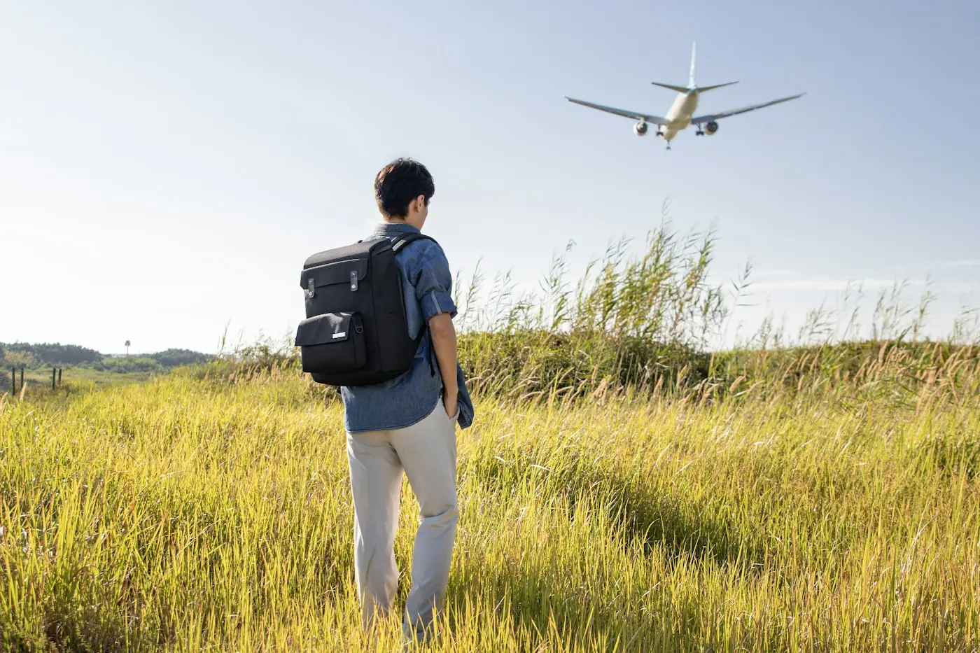 Man with a black backpack watching a plane fly low over a grass field