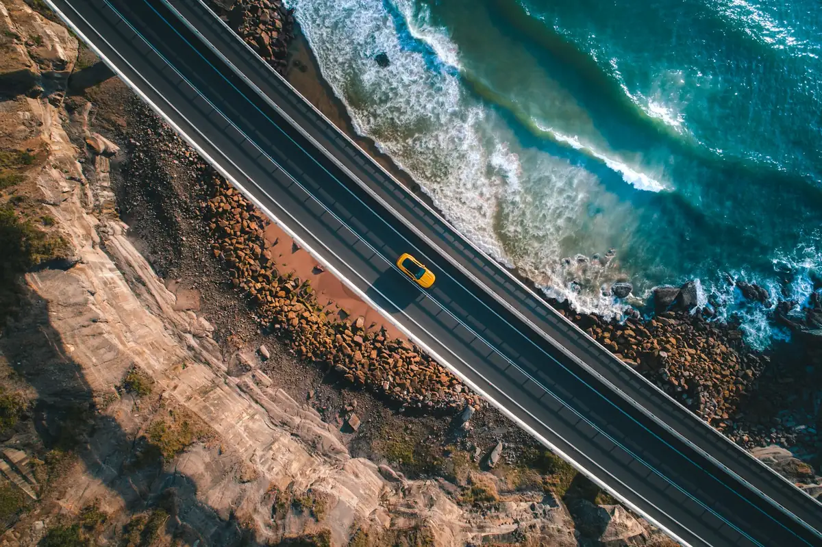 Aerial view of a small yellow car on a road that follows a rocky sea coast
