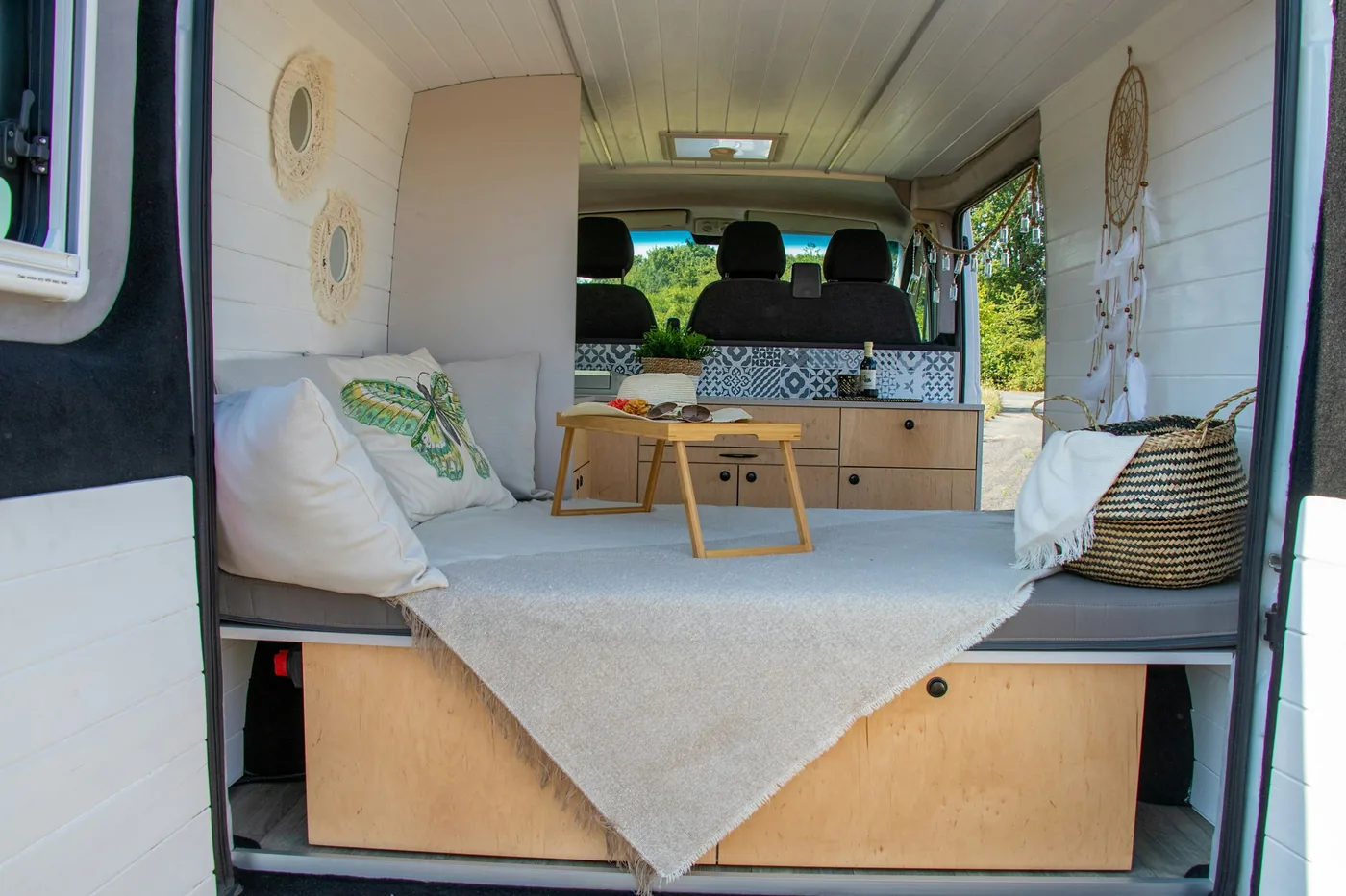Interior de una furgoneta reformada con una cama, cojines, una pequeña mesa de madera y una encimera