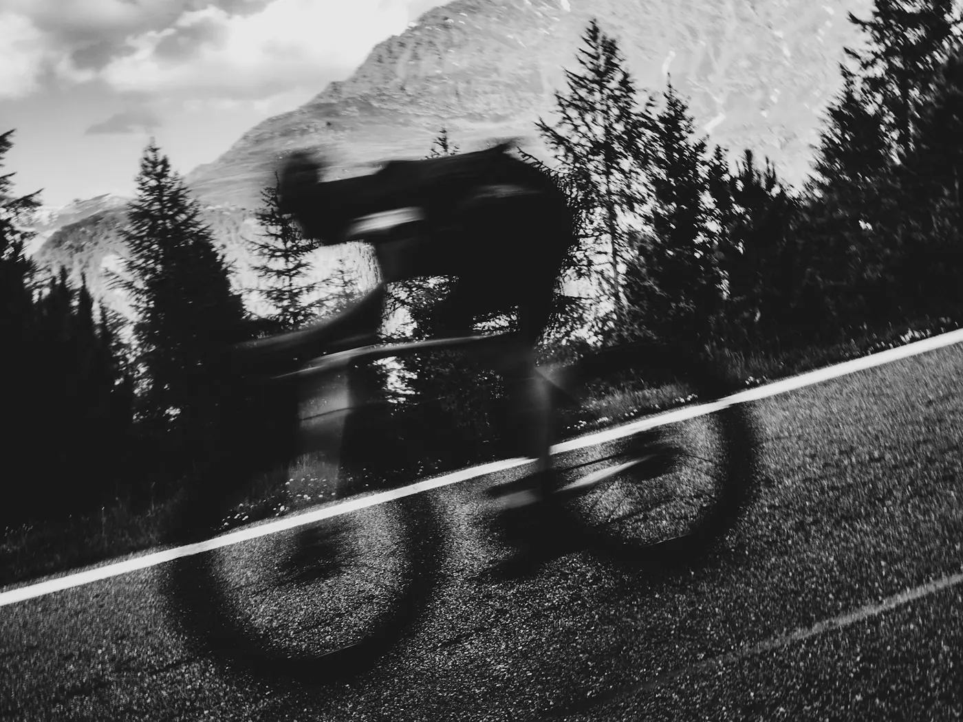 Schwarzweißfoto eines Radfahrers in Bewegungsunschärfe auf einer Bergstraße mit Kiefern und einem schneebedeckten Gipfel im Hintergrund