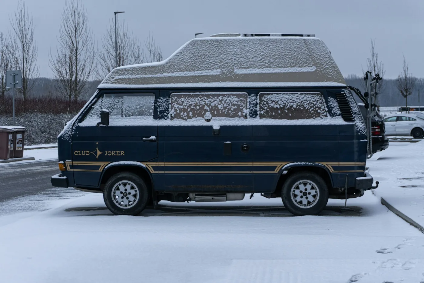 Dark blue camper van with a high roof covered in fresh snow, parked on a street