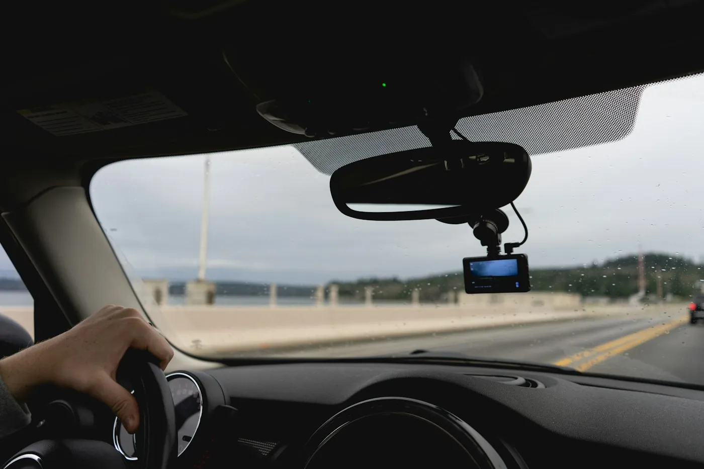 Dash cam mounted behind the rear-view mirror of a car on a highway, driver hand on the wheel