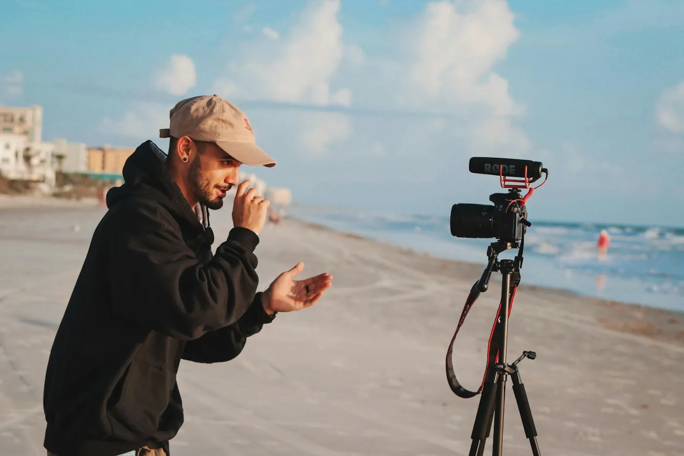 Man in a cap and black hoodie talking to a camera on a tripod on a beach, the kind of shot a wireless clip-on microphone is made for