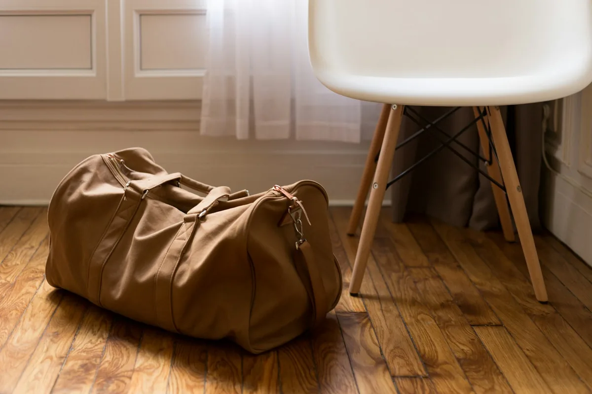 Een bruine duffel bag op een houten vloer naast een witte stoel