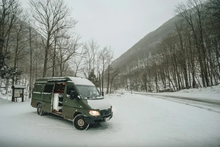 Green camper van with snow on the roof parked on a snowy road in a mountain valley with bare trees