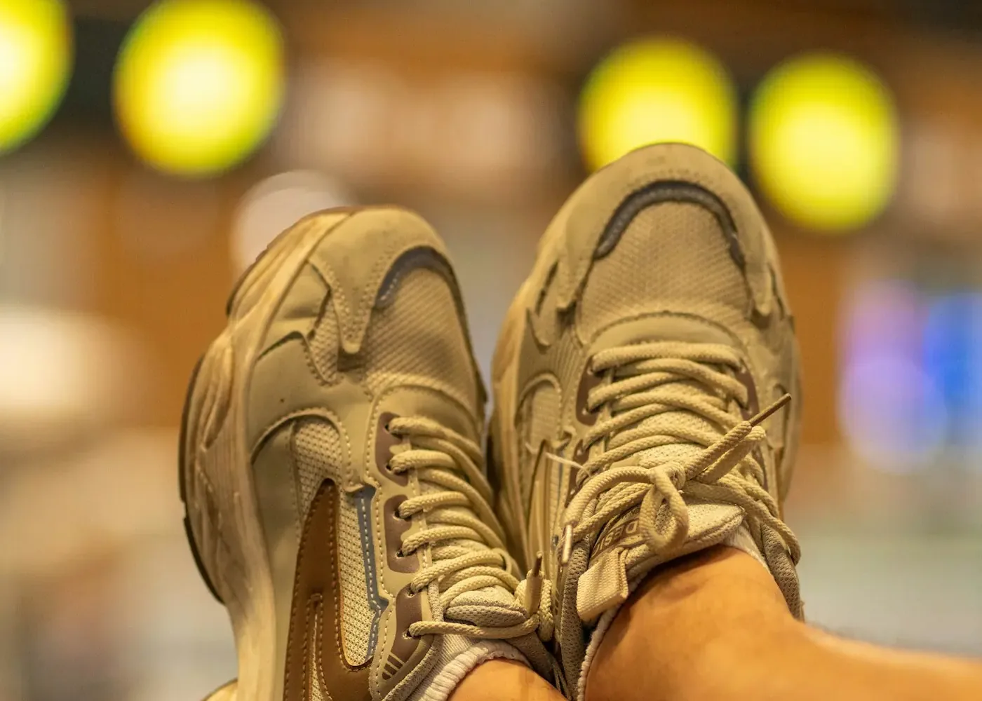 Feet in beige sneakers resting on a suitcase at an airport with blurred lights behind