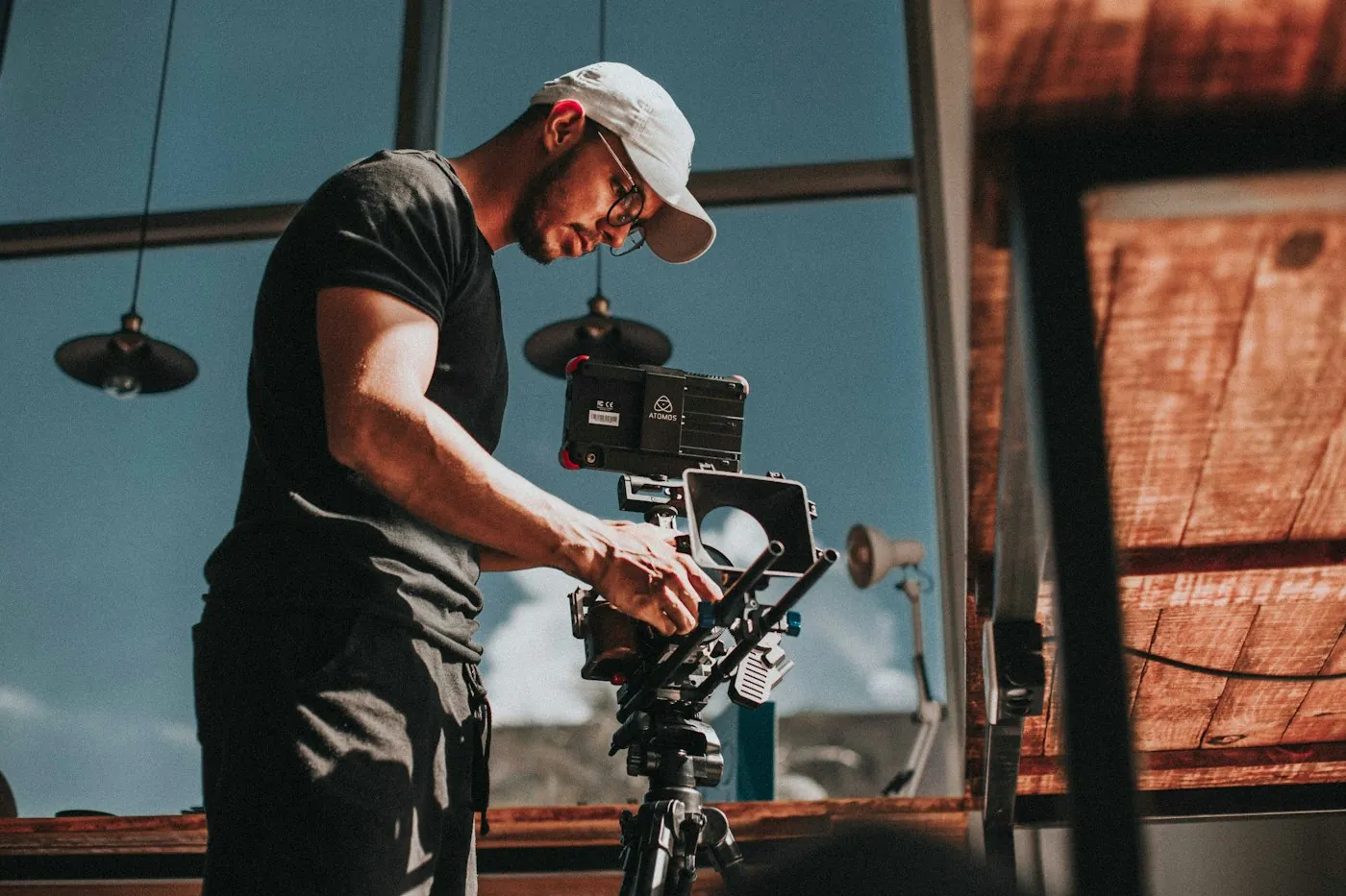Filmmaker in a cap and black t-shirt adjusting a camera on a tripod in a bright room