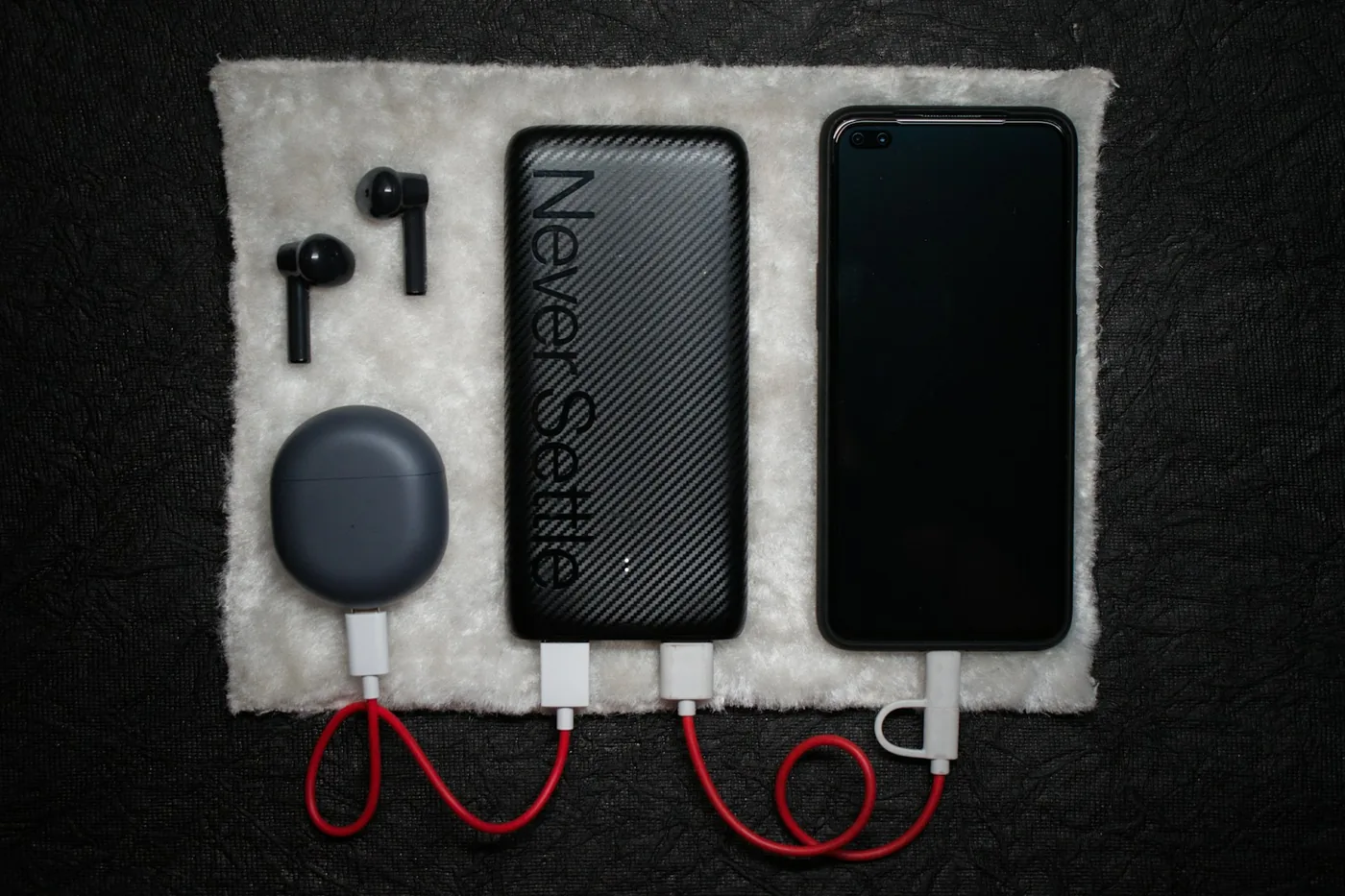 Flat Lay einer schwarzen Powerbank, eines Smartphones und In-Ear-Kopfhörern mit Ladecase auf weißem Fleece, verbunden durch rote Kabel