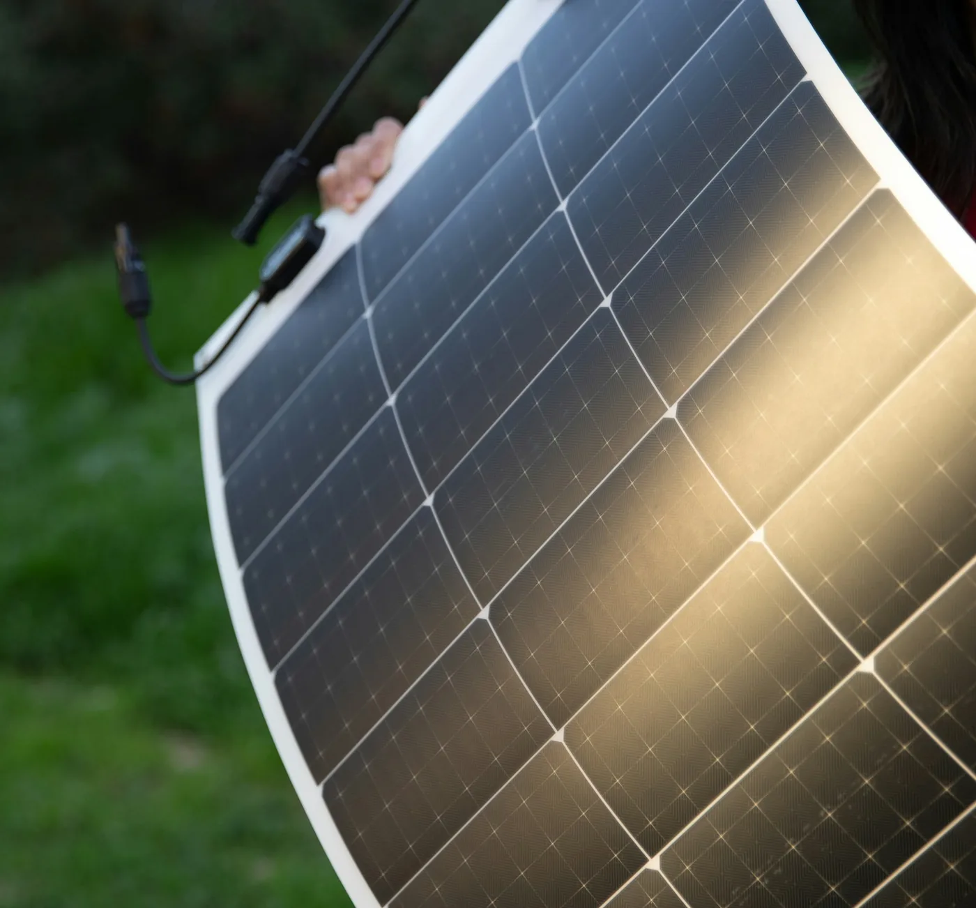 Hände halten ein dünnes, flexibles Solarpanel, das sich biegt, im Hintergrund ein grüner Hang