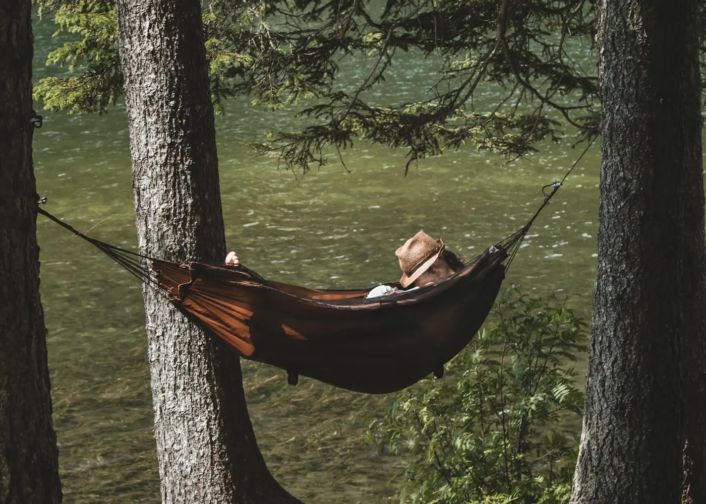 Person resting in a brown hammock between two trees by the water