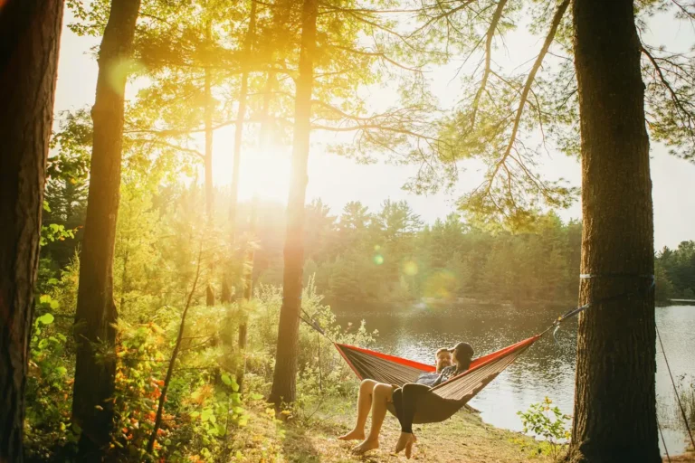 Two people resting in a hammock between trees by a lake at sunset