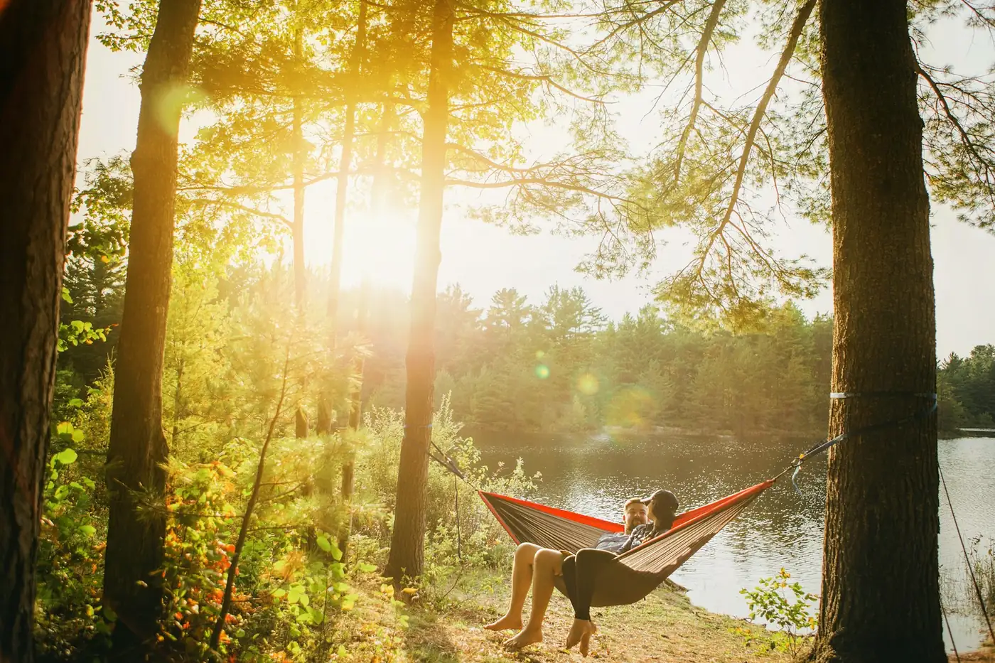 Two people resting in a hammock between trees by a lake at sunset
